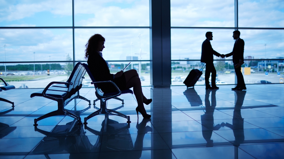 Flight Booking for Corporate Travel A Guide for Businesses Planning Trips to Brazil (1) Executiva usando tablet na área de embarque enquanto dois homens de negócios apertam as mãos - reserva de voos em grupo para empresas