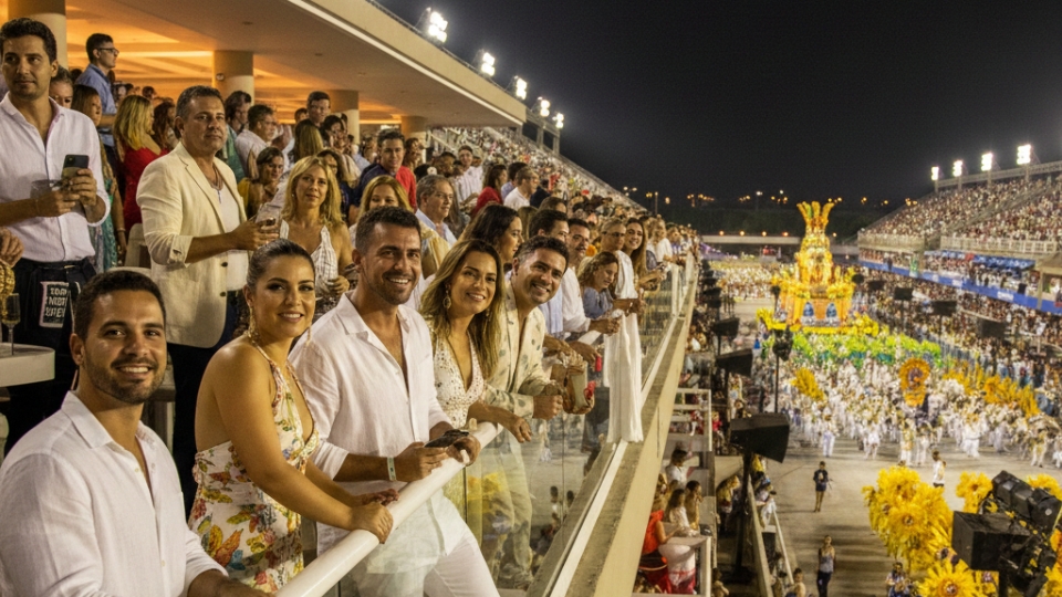 Pessoas aproveitando o desfile do Carnaval do Rio a partir de um camarote no Carnaval, usando roupas leves e assistindo às escolas de samba no Sambódromo à noite.