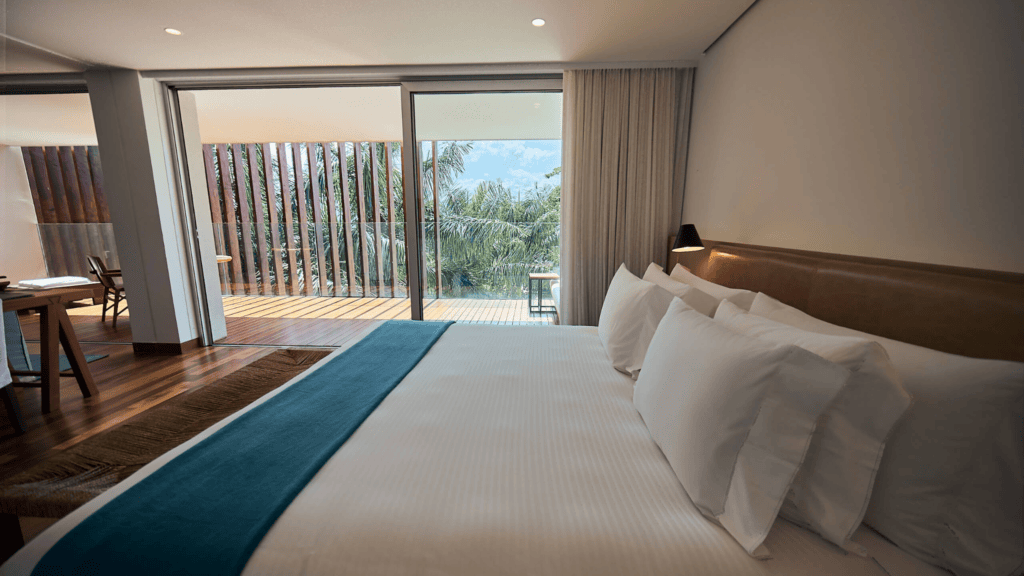 Fasano Angra dos Reis - Hotel Booking in Angra dos Reis (6) Modern hotel room with a large bed, balcony view of palm trees and ocean, and natural light.