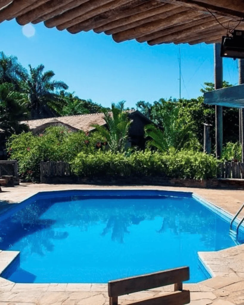 Pousada Aguapé - Hotel Booking in the pantanal Swimming pool with lush tropical trees and a thatched roof overhang in the background.