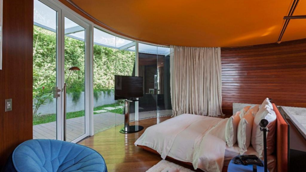 Unique Garden - Hotel Booking in Sao Paulo (9) Modern bedroom with large glass doors overlooking a garden, wooden wall, and a flat-screen TV.